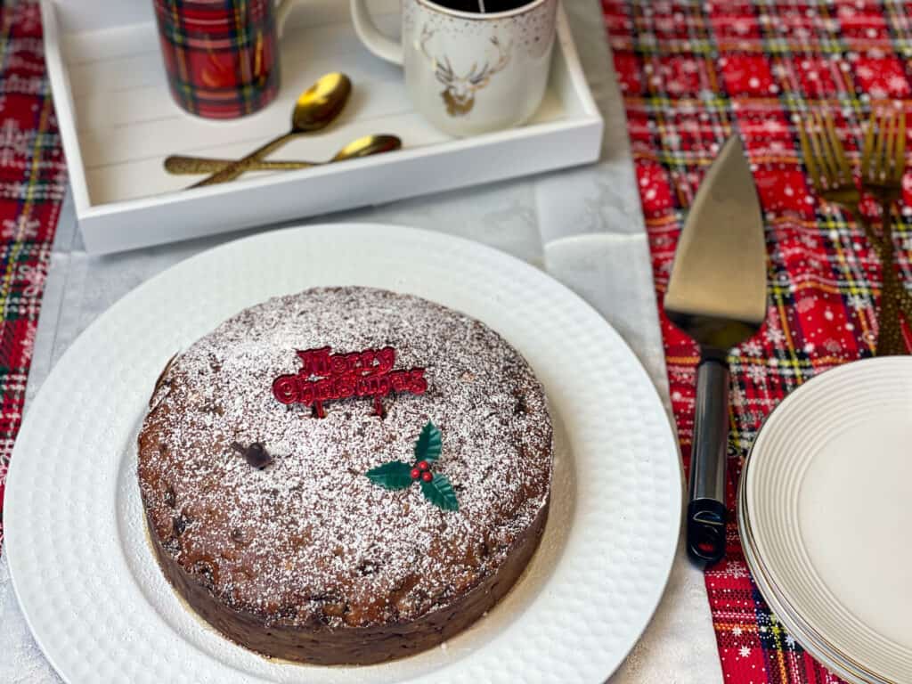 Christmas cake decorated and served on a white plate with tea tray in background and tartan table cloth.