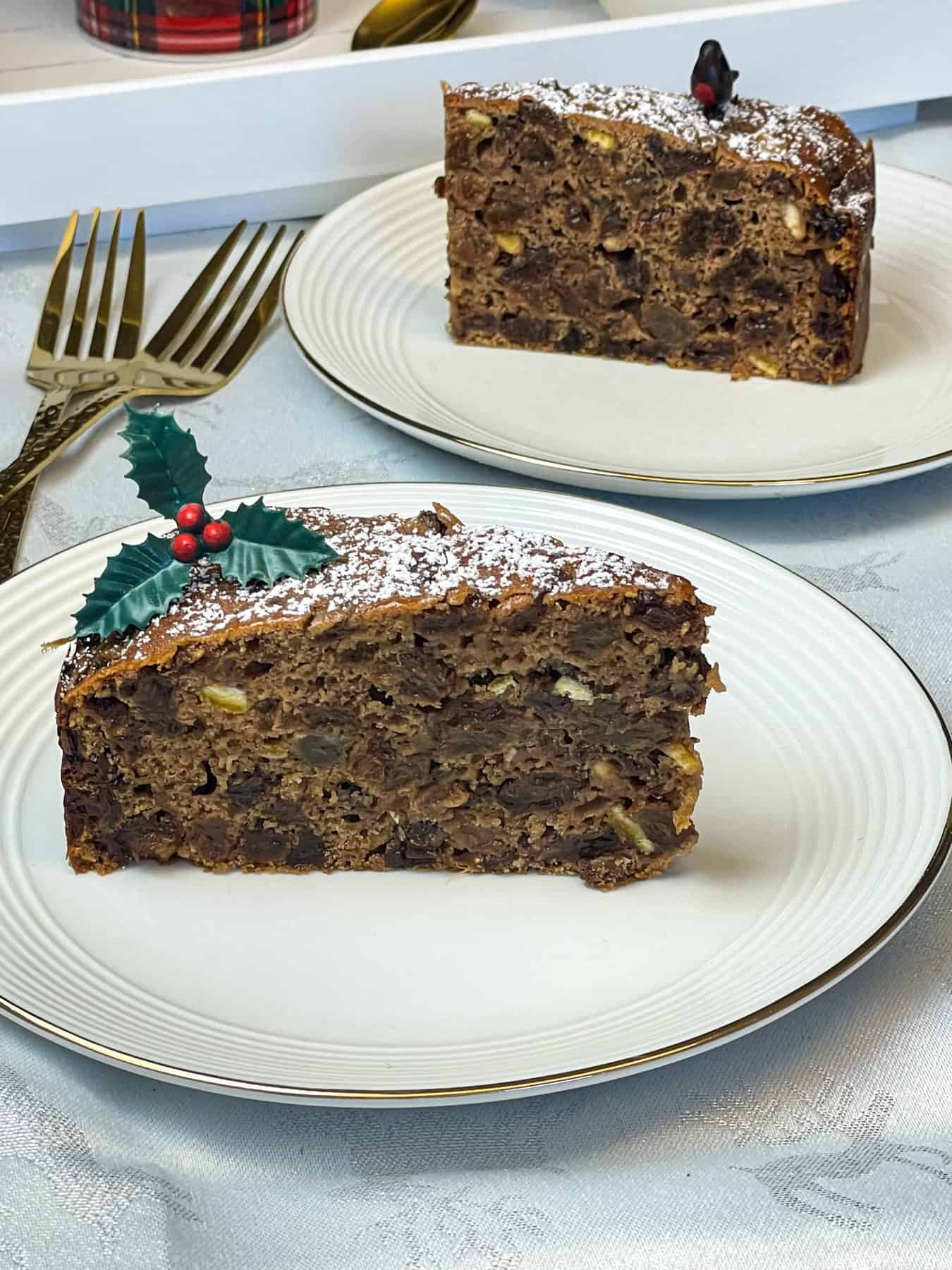 Two plates of sliced cake with a robin decoration and a holly decoration, with gold cake forks to side.