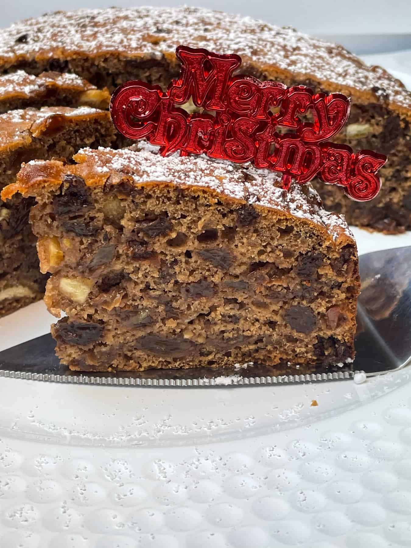 Slice of Christmas cake on serving plate with rest of cake in background.