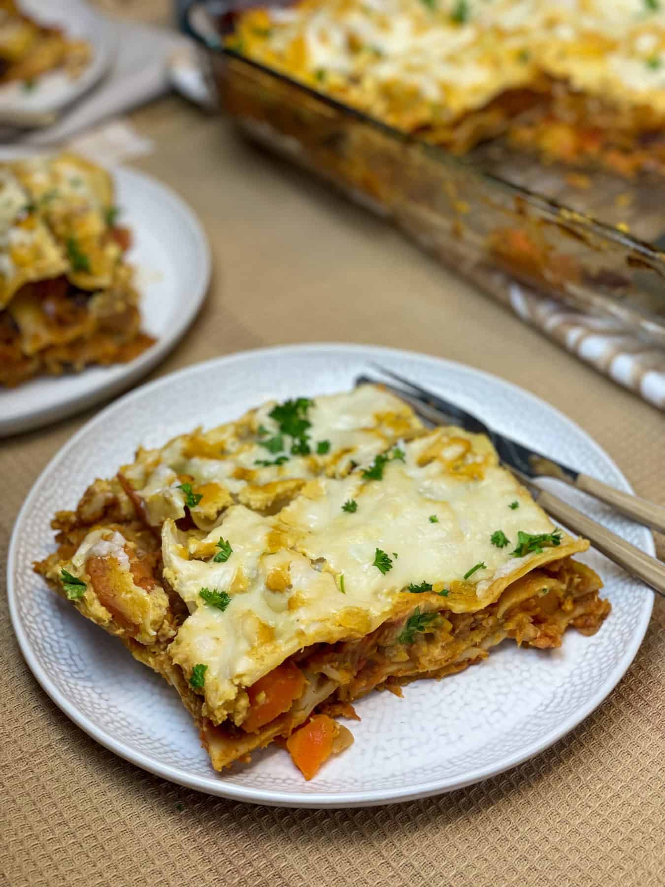 Plate with lasagna slice, fork and knife, second plate to side and baking dish to side, tan background.