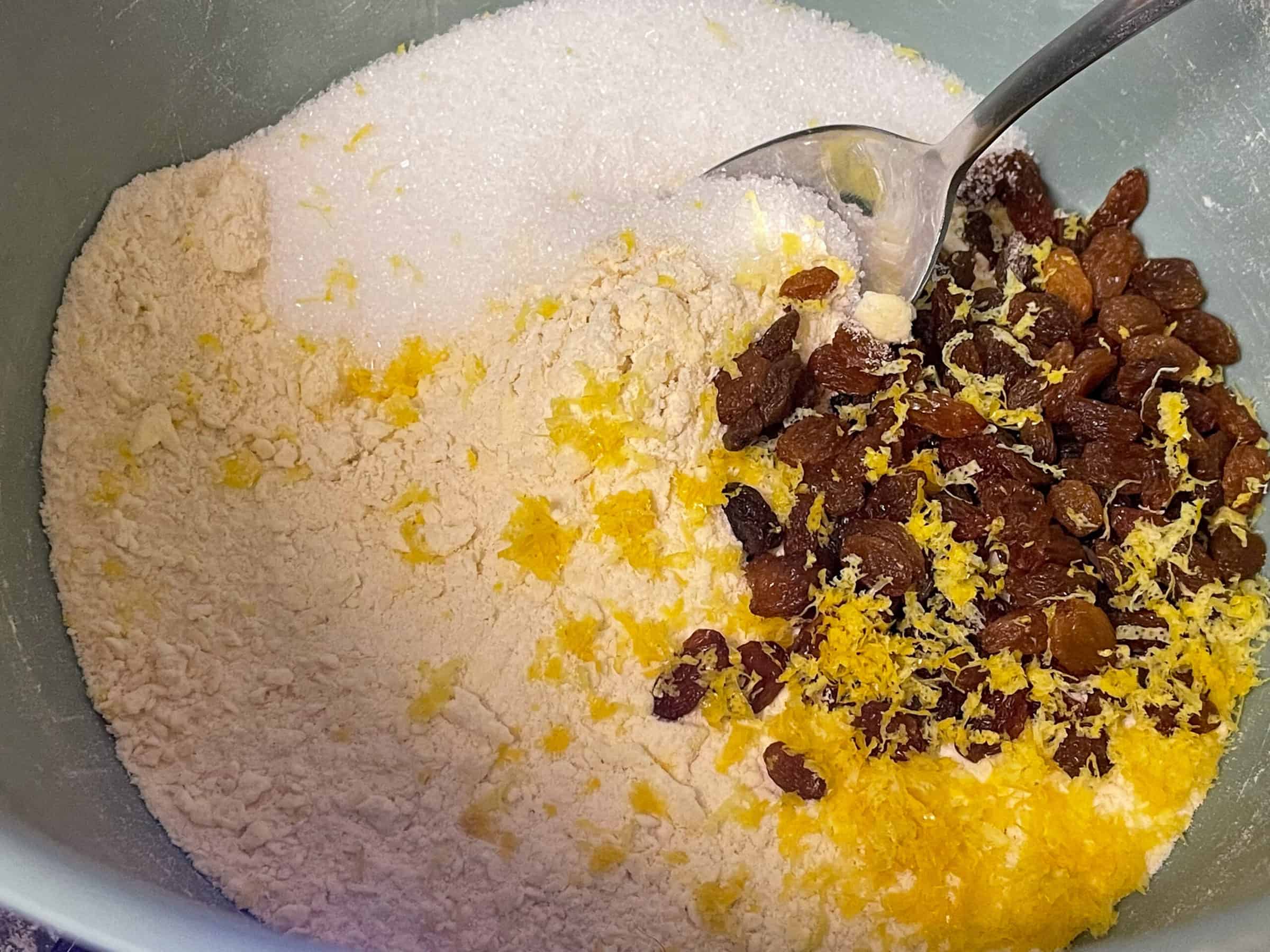 sultanas, lemon zest and sugar added to flour in mixing bowl, with silver spoon.