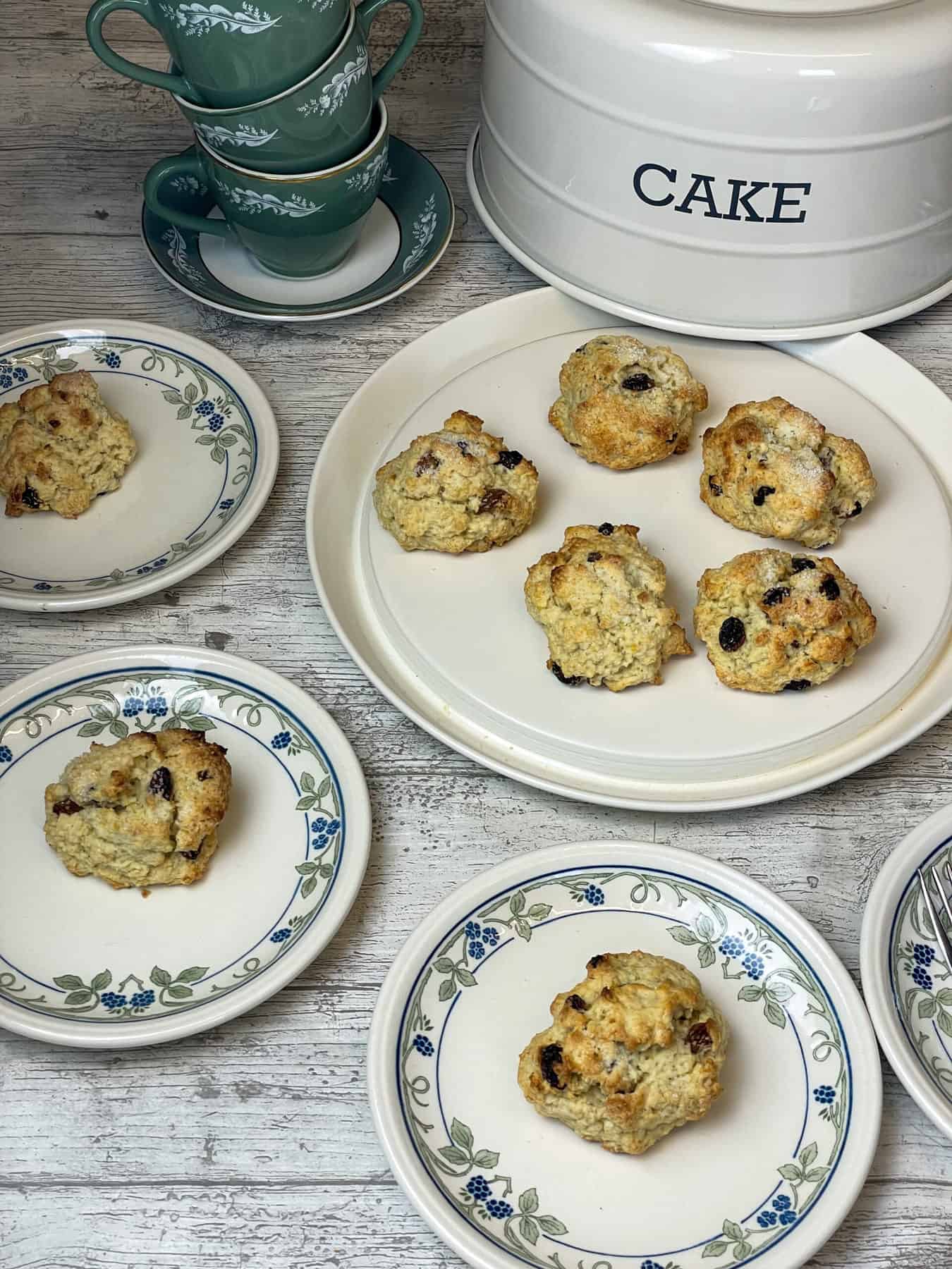 Rock cakes on a tea table on small plates with cake tin in background, green tea cups to side.