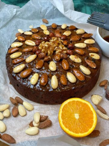 Vegan quick and easy Christmas cake decorated with whole nuts, nuts to side and sliced orange to side, greaseproof paper background and green sparkle backdrop, featured image.