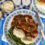 A blue and white patterned plate with vegan coq au vin served with mashed potatoes and green beans, bread rolls to side with salt and pepper shakers.