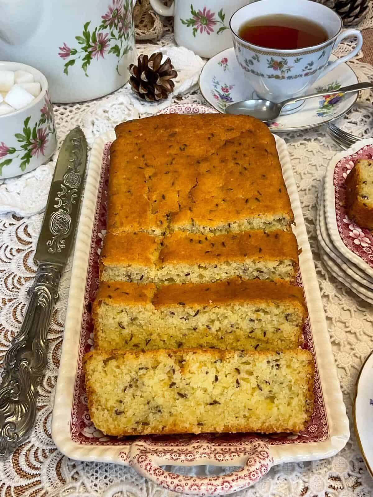 Vegan seed cake on a vintage tray with pink flower pattern, antique cake knife to side, flower patterned teapot, sugar dish, milk jug, and tea cup and saucer to side, crochet tablecloth background.