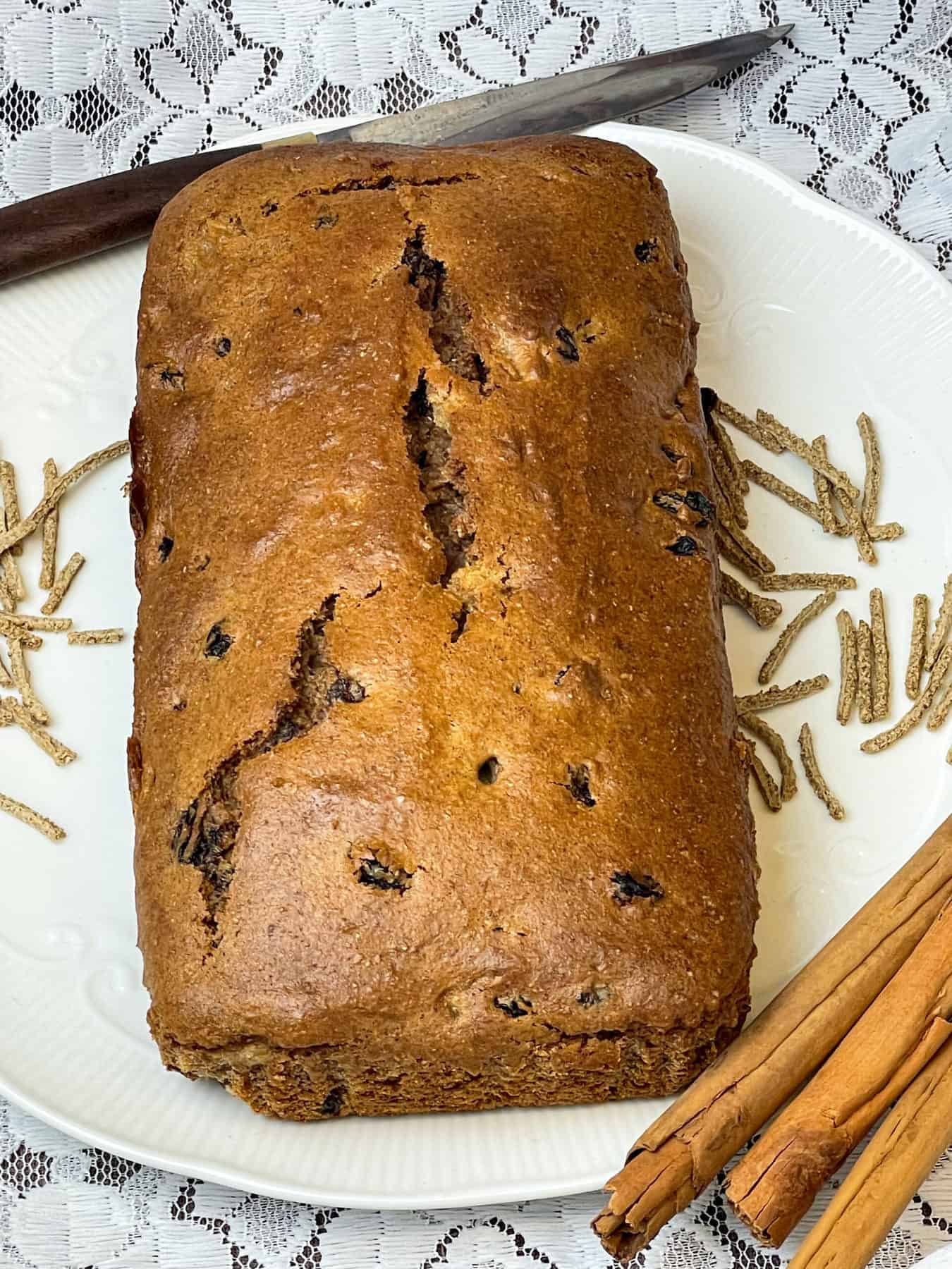 all bran cinnamon loaf on white plate with wooden handled knife, sprinkle of all bran cereal, sticks of cinnamon, white doily background. Featured image.