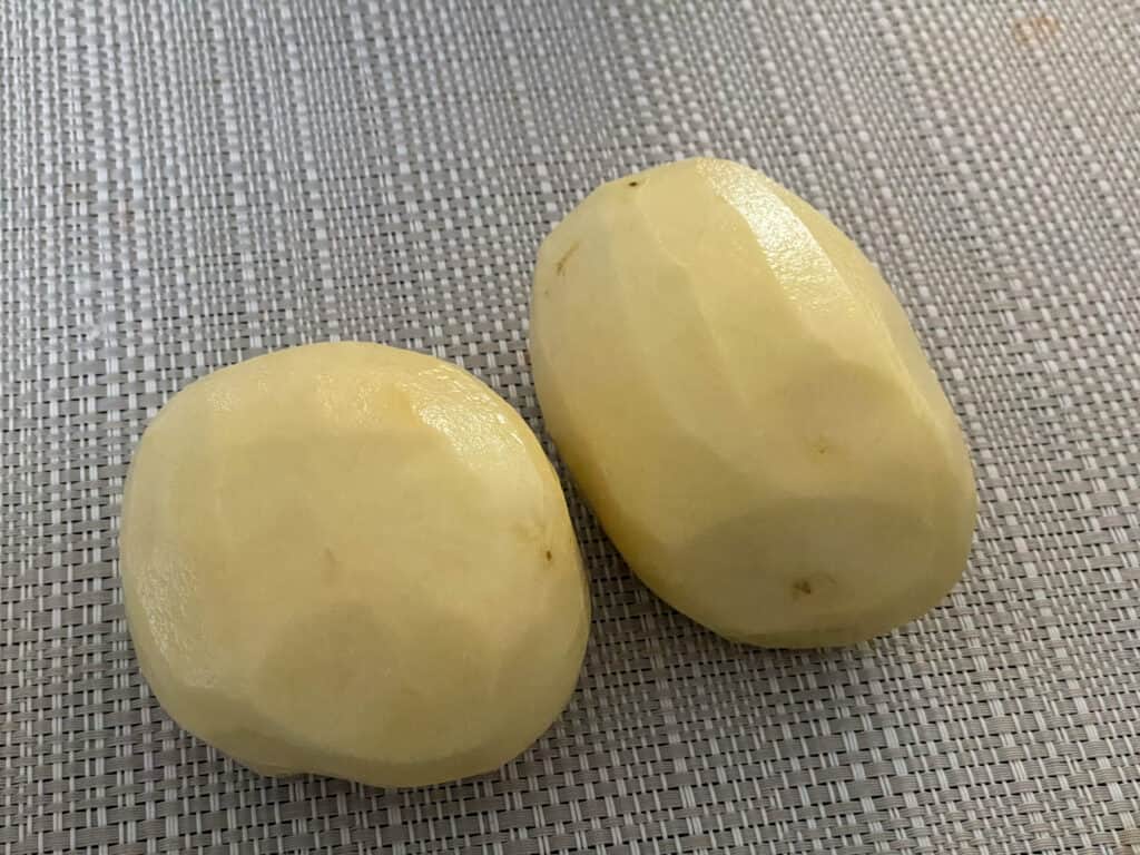 Two peeled potatoes on creamy white placemat.