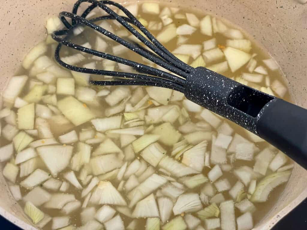 Chopped onions added to veggie stock with a whisk in the creamy saucepan.