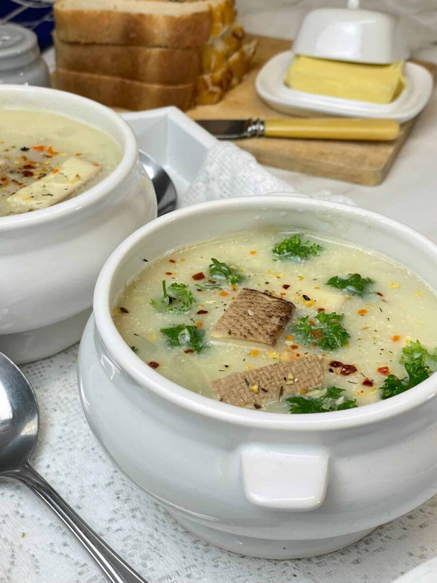 Close up of small soup tureen filled with vegan Scottish Cullen skink soup and second bowl to side with silver spoon to side sitting on a white doiley placemat, mini butter dish with slices of bread in distance, featured image.