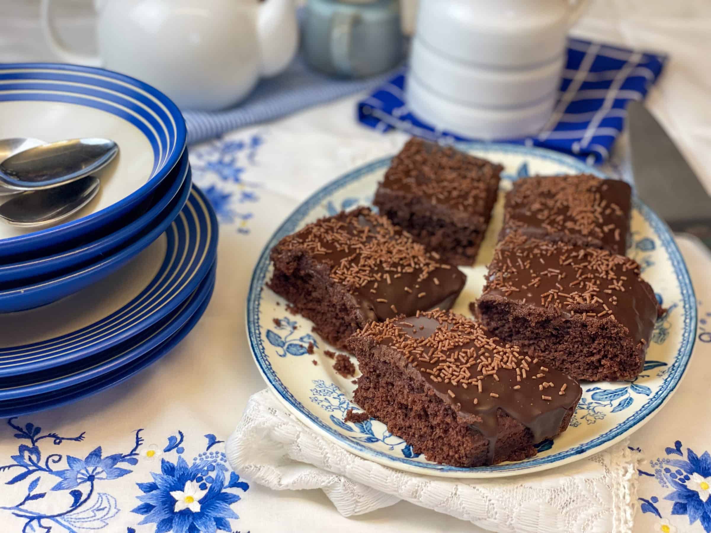 Oval cake plate with slices of chocolate sponge pudding, custard jug to side and serving bowls and plates with blue rim to side, white teapot and small blue mug in distance, blue flower white table cloth.