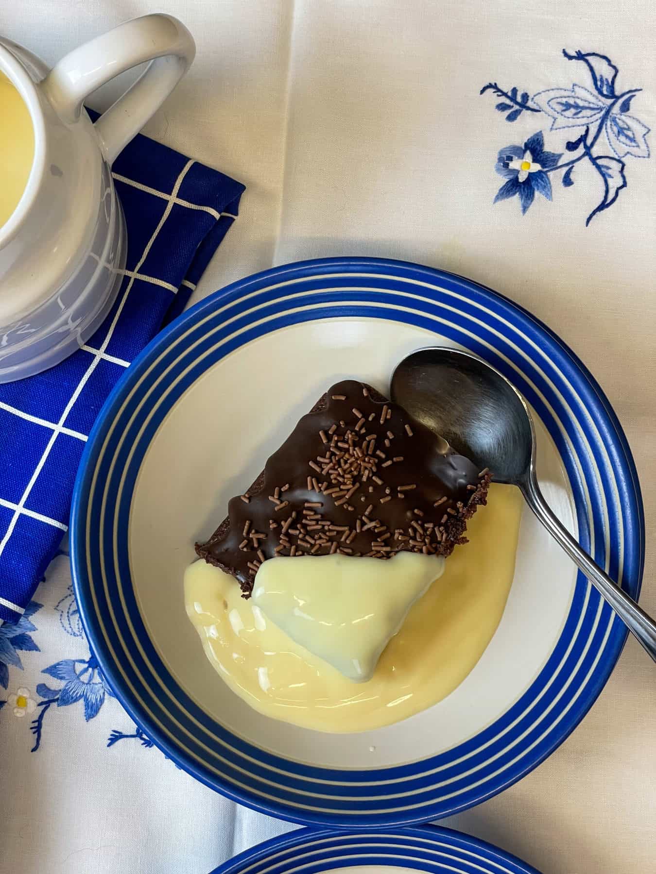 A blue rimmed white bowl with a slice of chocolate sponge with custard poured over, silver spoon to side, custard jug to side, and blue flower table cloth background.