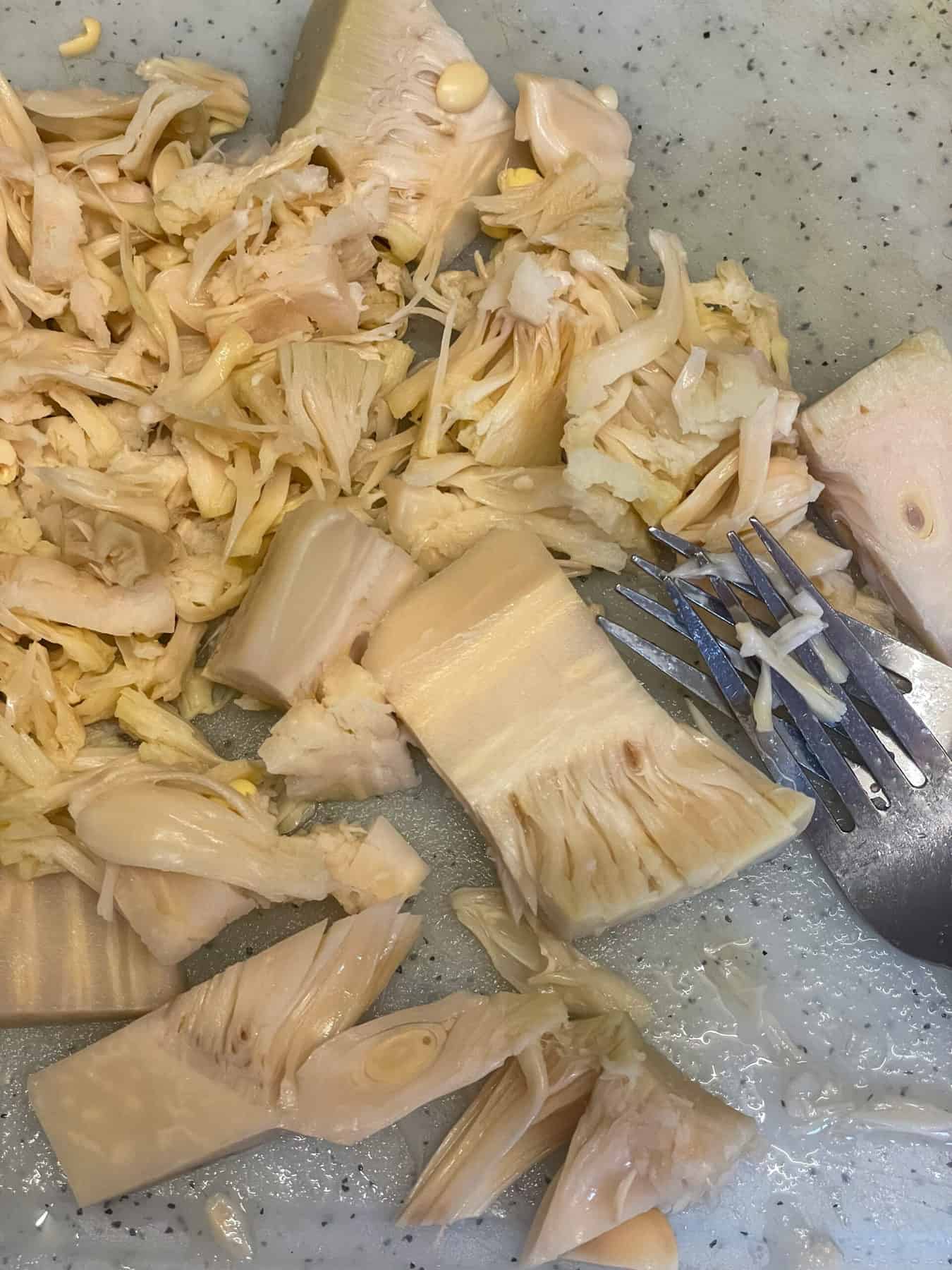 Jackfruit on a white chopping board being shredded into little pieces with a silver fork.