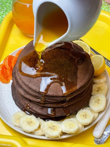 Vegan chocolate pancakes with a jug drizzling syrup over the pancake stack, sliced bananas and orange segments around the serving plate, glass of orange juice to side, yellow serving tray, featured image.