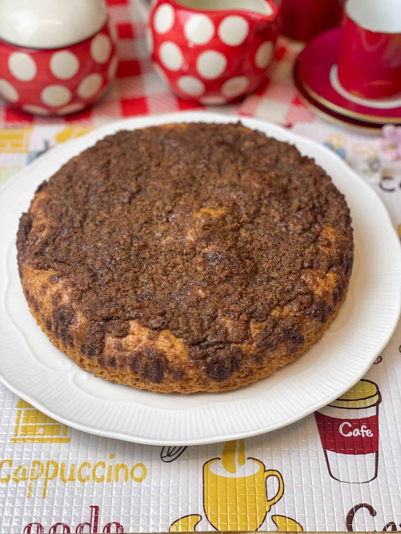 whole coffee cake on white cake plate with coffee patterned table mat and red spotty sugar bowl, red spotty milk jug and small coffee mug and saucer to side.