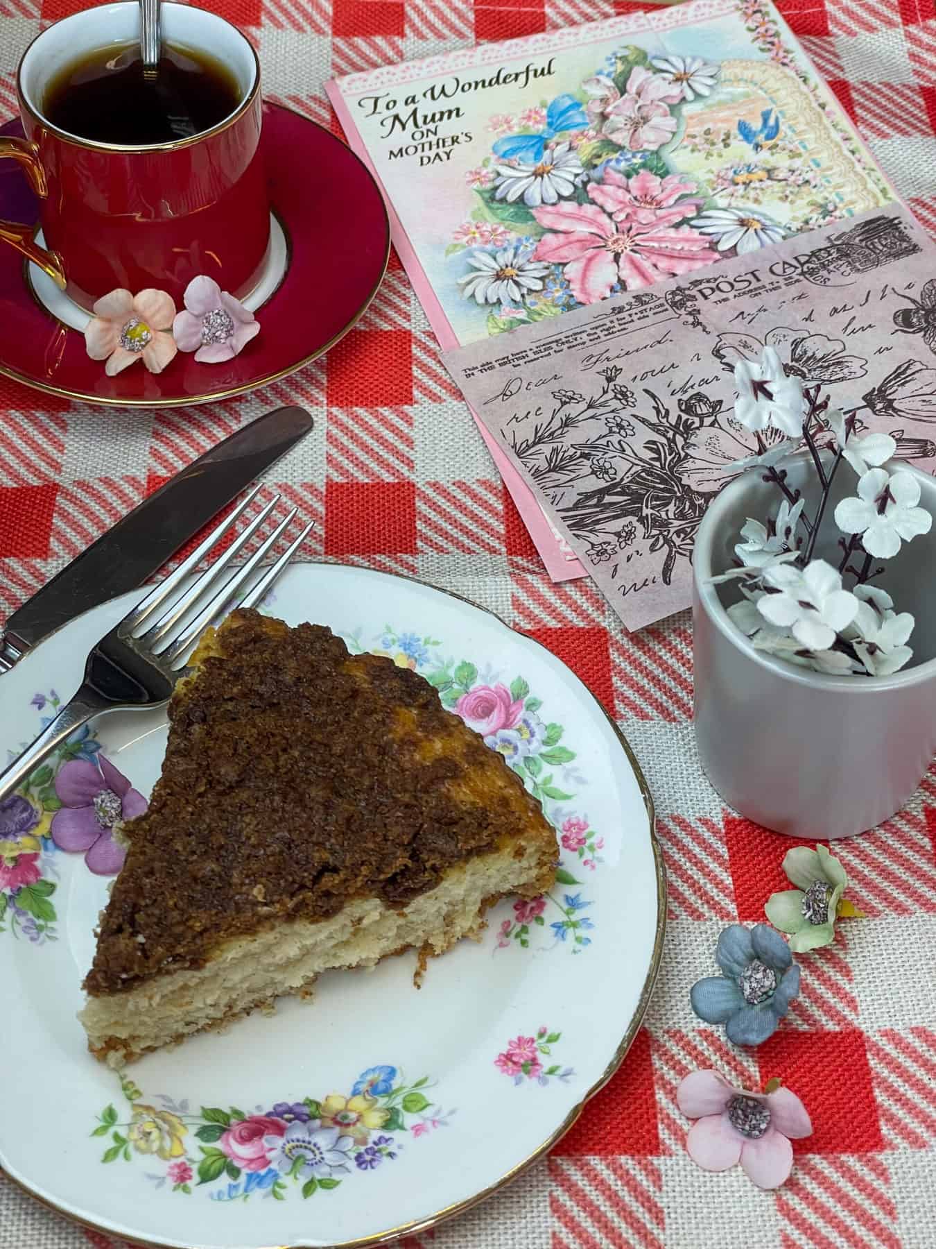 Slice of coffee cake on flower plate with cake knife and fork, small red coffee mug and saucer to side, Mother's day card to side and small grey mug with white flowers to side, red and white check tablecloth, small flower heads scattered around.