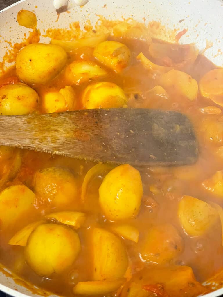 stock added to potatoes cooking in curry spiced gravy in pan, being stirred with wooden spatula.