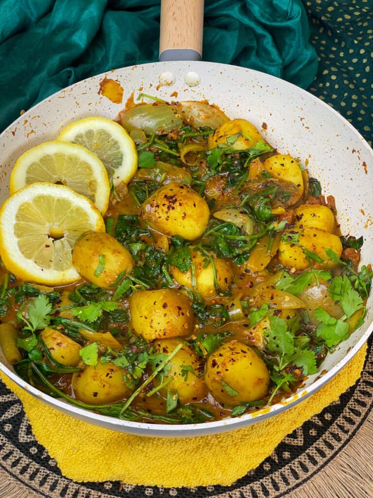 Saag Aloo with herb garnish, three lemon slices and red chilli flakes, pan on yellow tea towel, and tan fringed mat, green background.