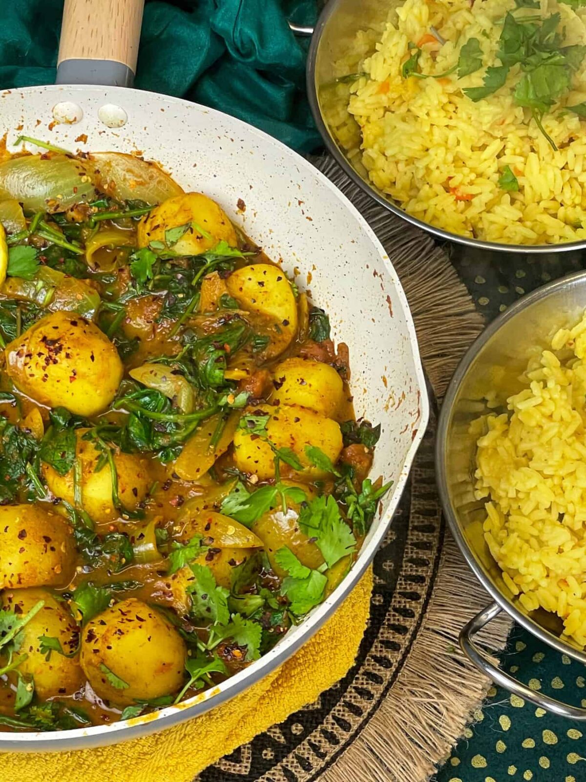 side view of pan with saag aloo with coriander garnish, 2 Balti dishes of yellow rice to side, yellow mat under pan, brown ruffled mat and green background.