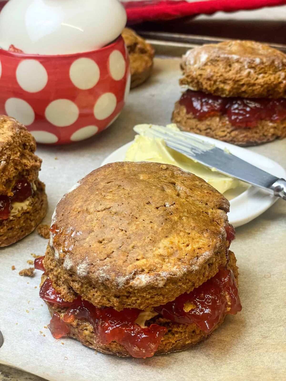 A large scone on baking tray with jam filling, butter dish to background along with red and white spotty jam jar, two treacle scones to side also.