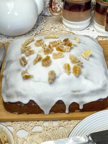 iced gingerbread cake on wooden chopping board with pieces of crystalized ginger on top, brown mugs to background along with a white tea pot.