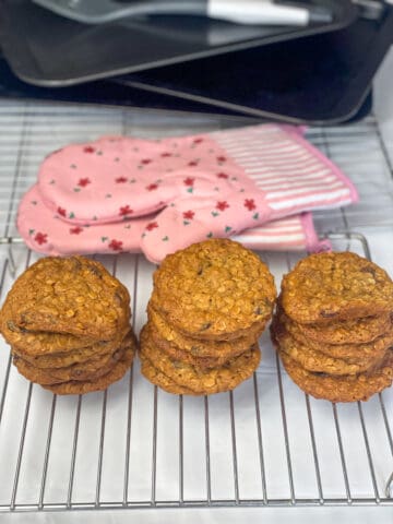 Three piles of oatmeal raisin cookies on wire rack with two pink flower oven mitts in background, with pile of baking trays and spatula in far background.