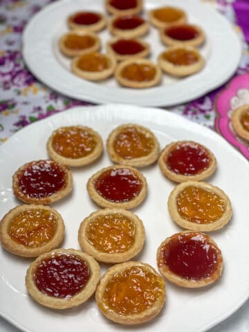 Two white plates filled with jam and marmalade tarts with purple flower background, small serving plate to side with pink flower rim.