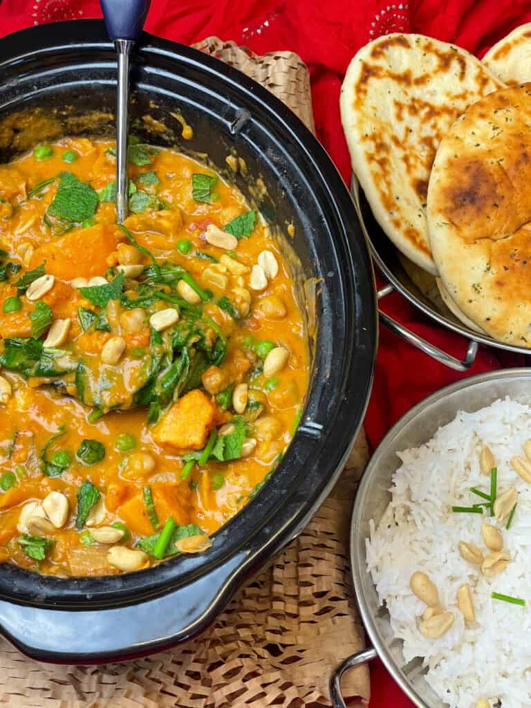 Thai curry in slow cooker garnished with peanuts and fresh herbs, silver Balti dish with rice to side, and Balti dish with naan breads to side, red background and paper raffia under slow cooker, serving ladle in curry.