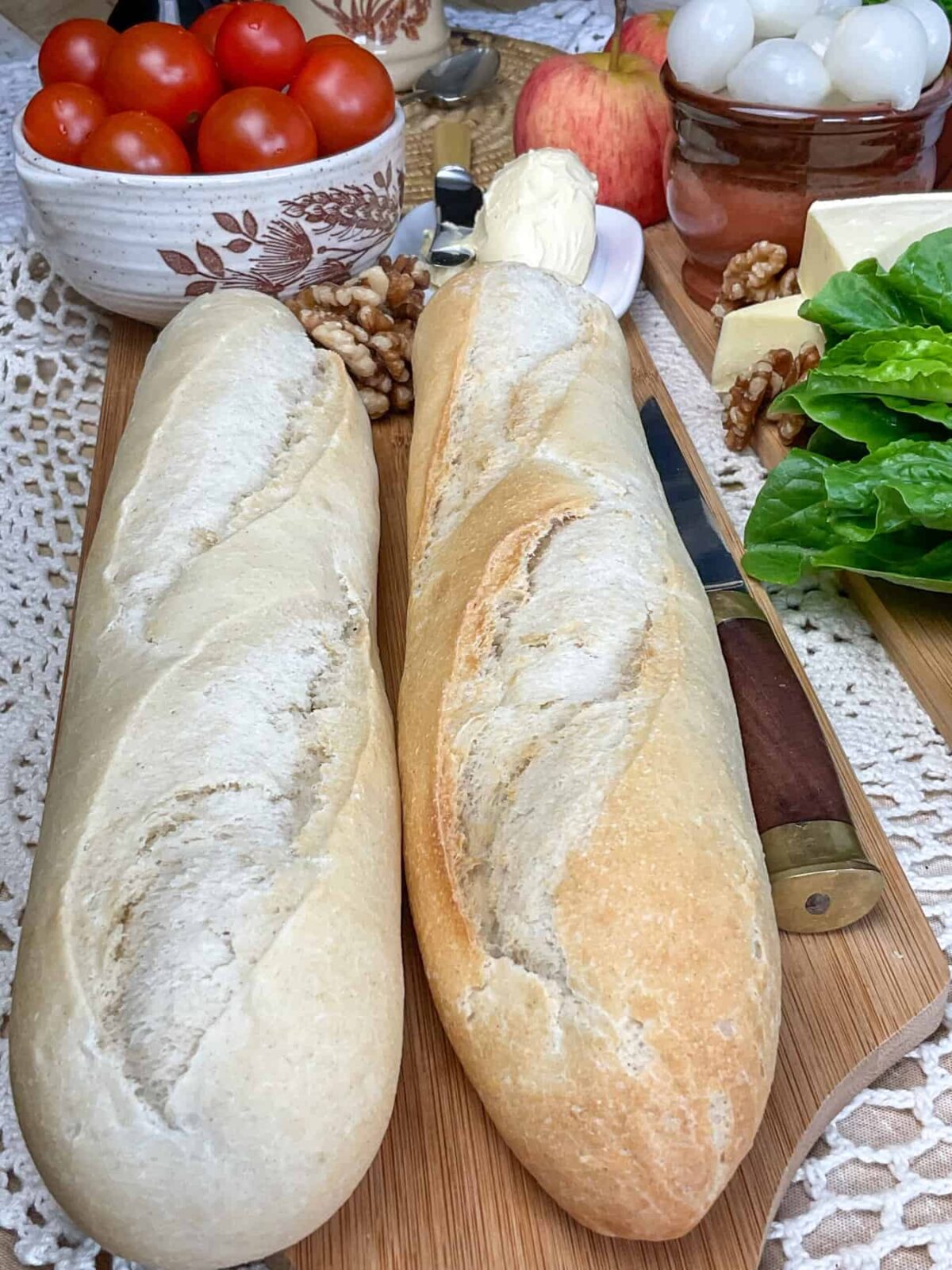 Two unfilled baguettes sitting on cutting board with bowl of cherry tomatoes to side and lettuce to side.