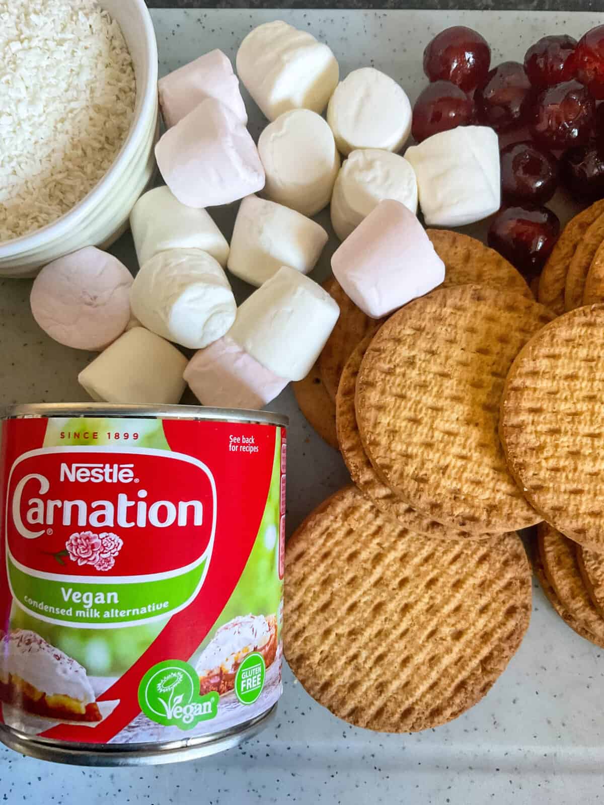 white board with vegan condensed milk can, digestive biscuits, marshmallows, candied cherries and small bowl of desiccated coconut.