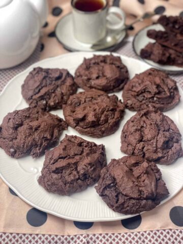 A plate filled with double chocolate chip cookies with white teapot and cup and saucer in background.