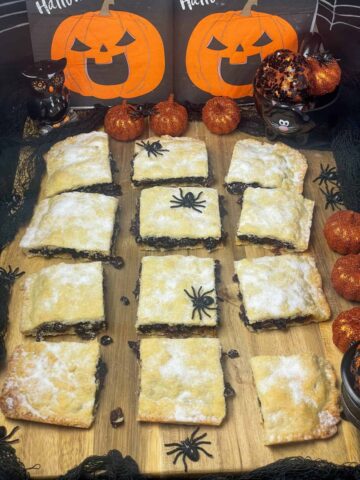 Fruit slice on wooden board with Halloween decor including fake spiders, pumpkin napkins, pumpkin ornaments, owl salt shaker, and a black cat cup measurer, black net around the board.