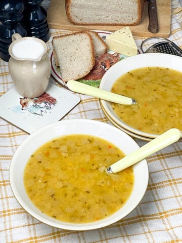 two bowls of yellow split pea soup on a yellow and white striped table cloth, cream jug to side, bread with slices on wooden board, black salt and pepper shakers, and a piece of vegan cheese.