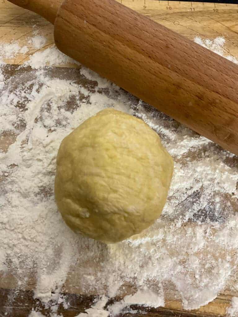 Pastry dough ball on floured chopping board with wooden rolling pin to side.