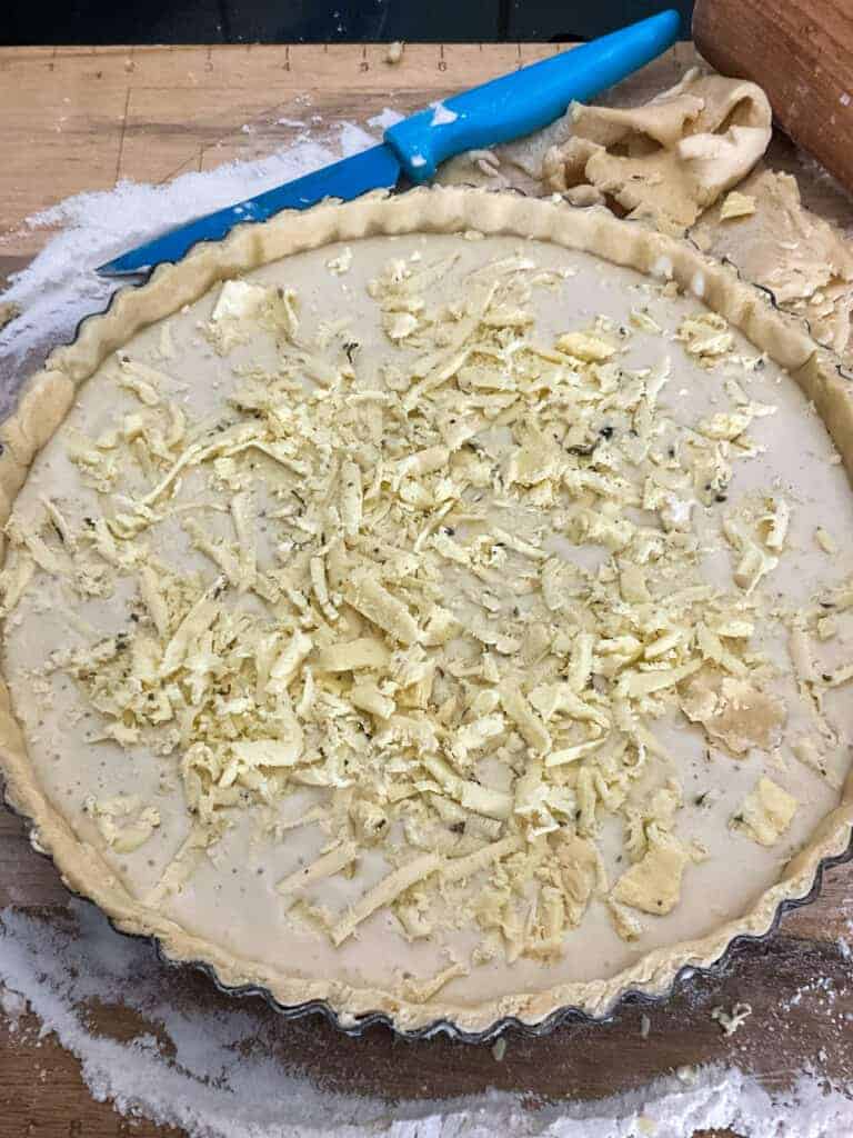 Grated vegan cheese added to unbaked cheese flan on chopping board.