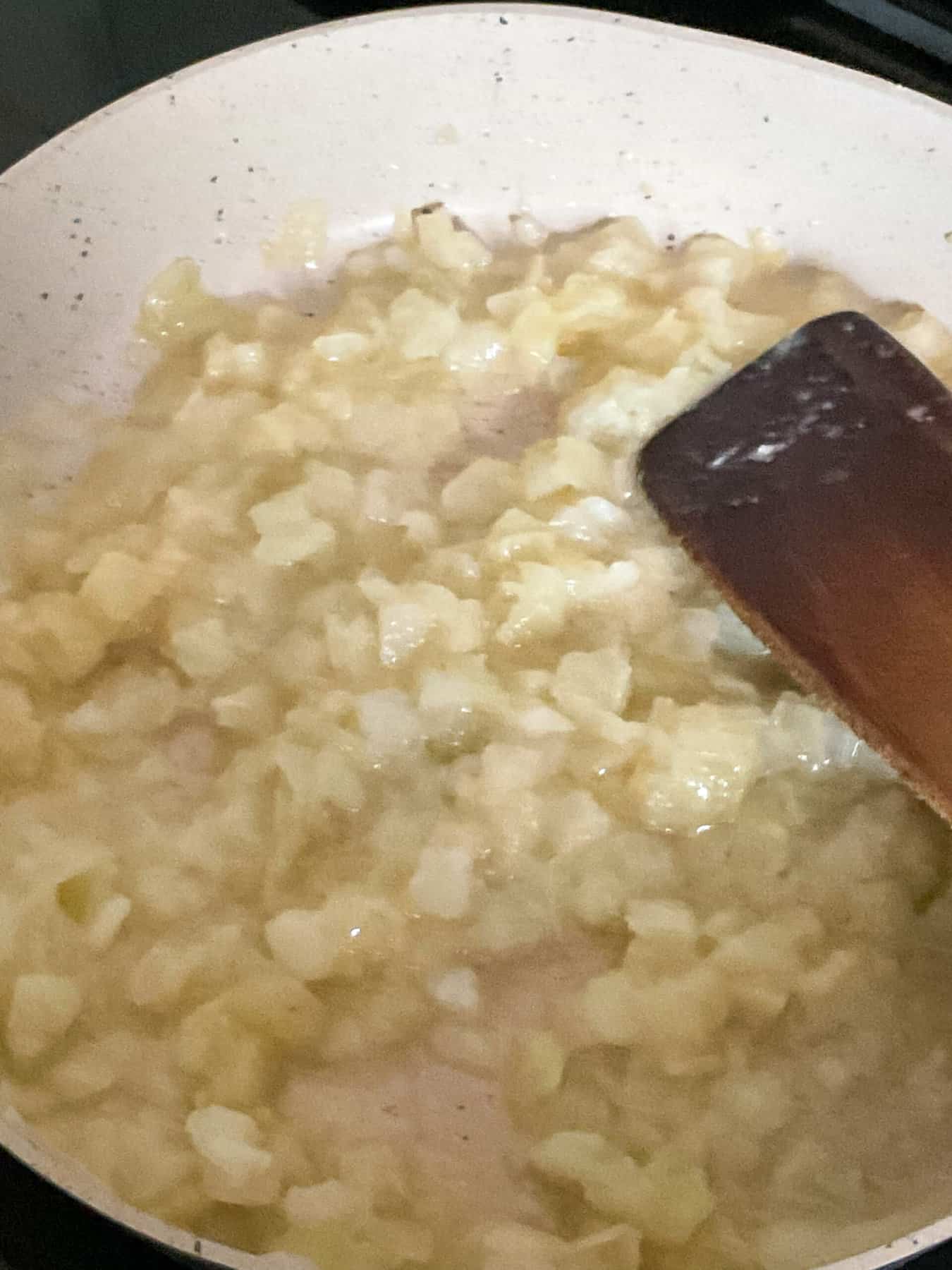 Onions cooking in coconut oil in cream coloured fry pan with wooden spatula.