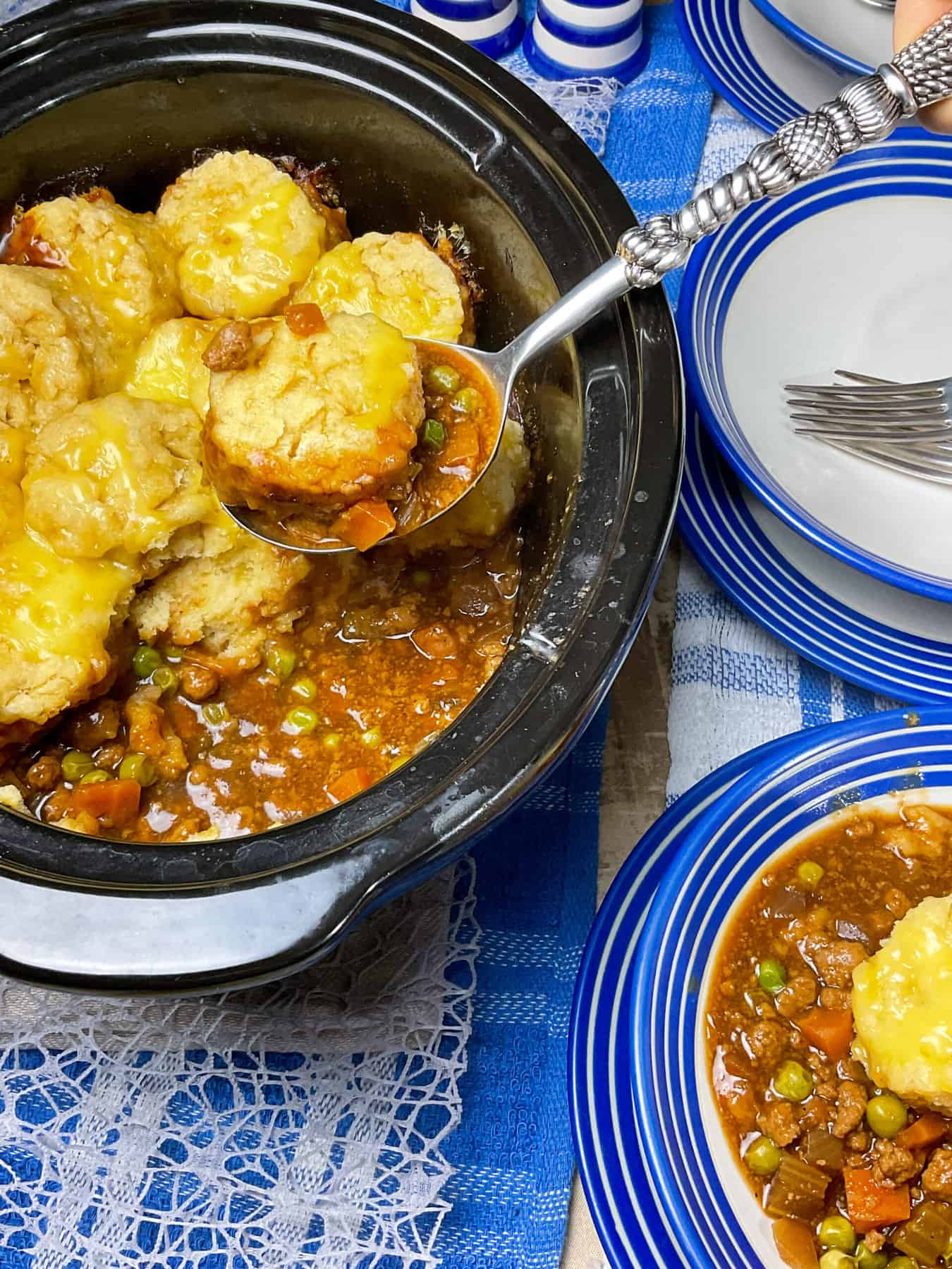 slow cooker pot with cobbler inside and silver serving spoon scooping a portion up, three blue rimmed bowls to the side with one with a serving of cobbler, blue and white table cloth background.