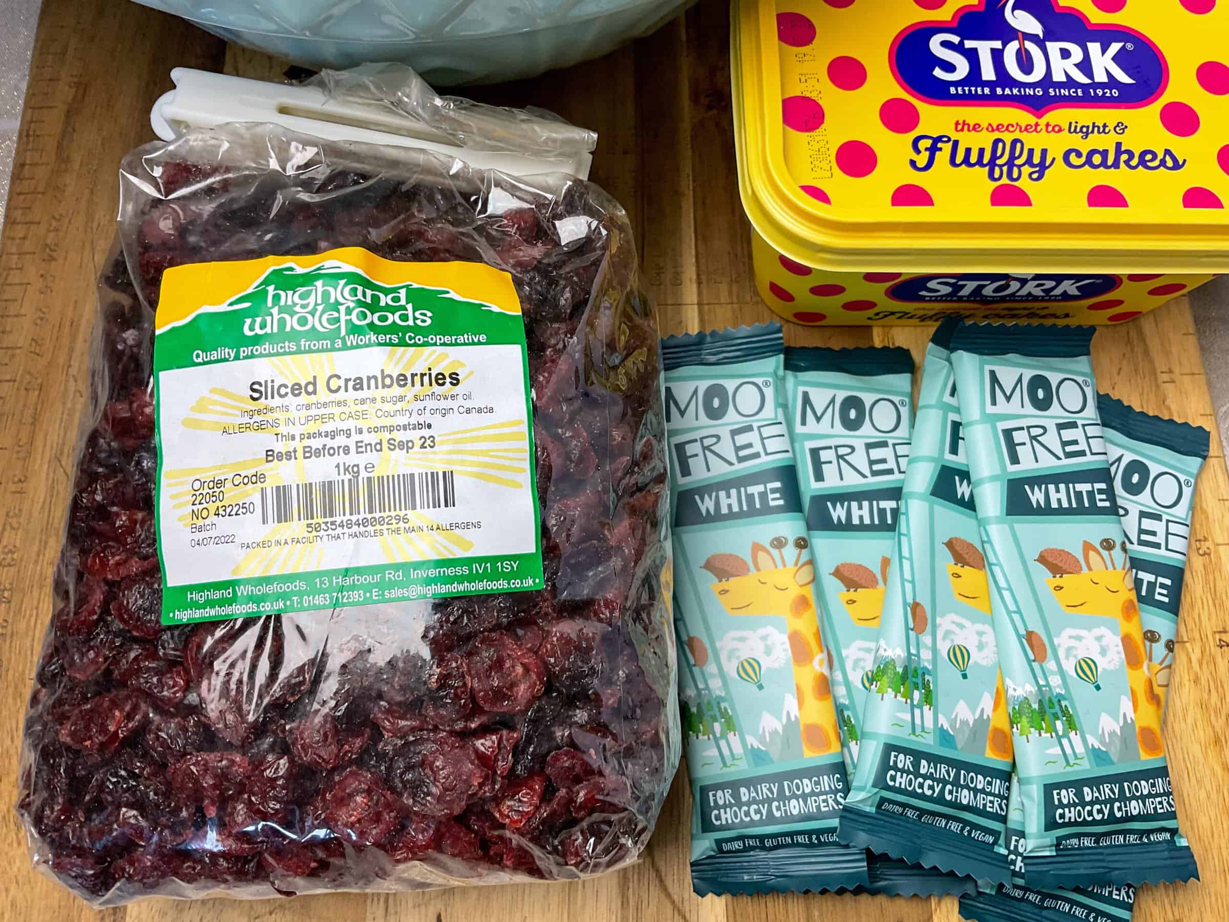 dried cranberries in a package, five small bars of Moo Free white chocolate bars and a tub of stork margarine on a wooden chopping board.