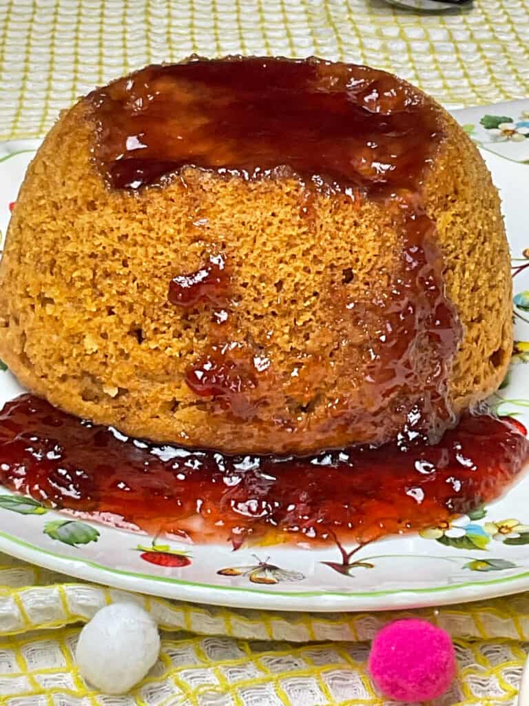 vegan steamed jam pudding on serving plate with yellow and white tea towel with little pom-pom decor.