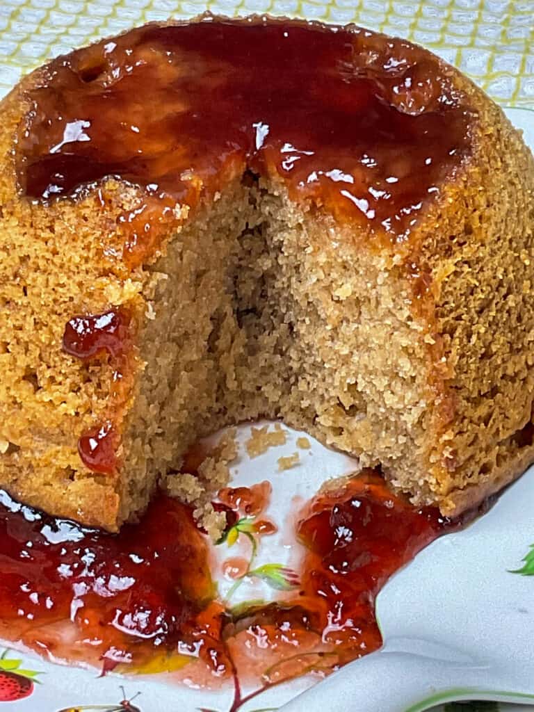 A close up of steamed jam pudding with a slice removed and jam dripping down onto the fruit and flower patterned plate.
