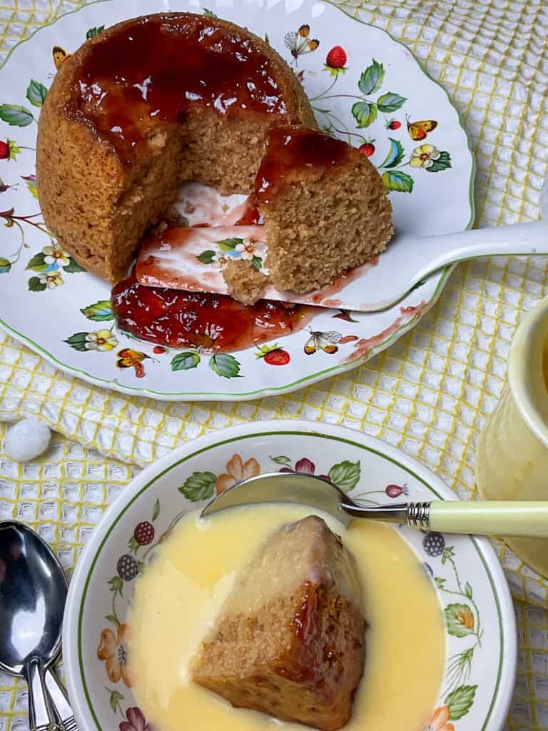 steamed jam pudding on flower and fruit image cake plate with matching spatula to background, slice of pudding in flower rimmed bowl with custard, yellow jug to side, and yellow tea towel background.