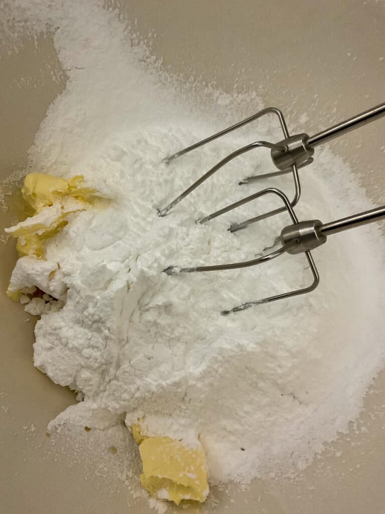 cinnamon butter cream frosting ingredients added to mixing bowl with electric hand mixer blades in the bowl.