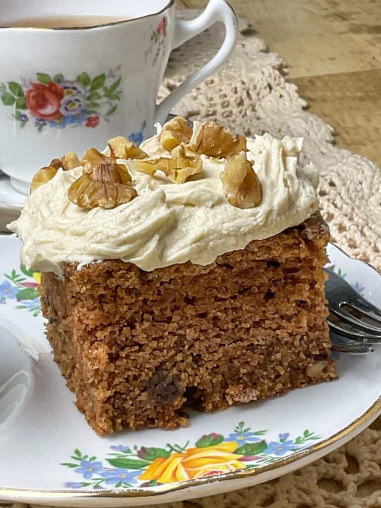 applesauce cake decorated with cinnamon buttercream frosting and on cake plate with flower image and china tea cup with flowers in background.