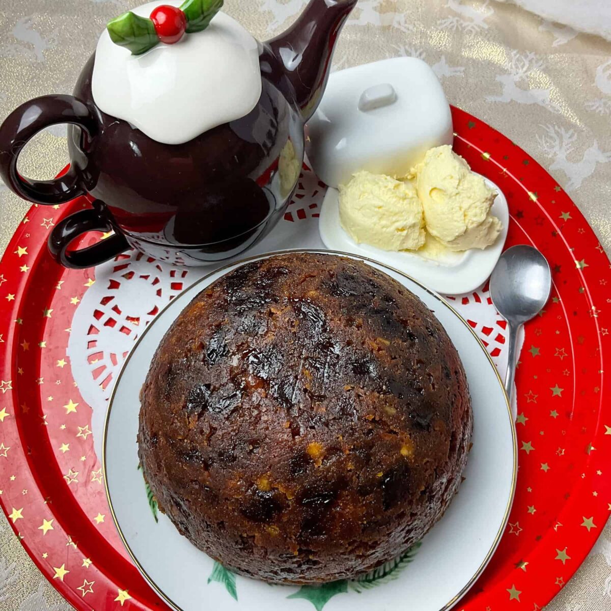 Christmas pudding on serving plate, with teapot to side that resembles a Christmas pudding with holly on top, brandy butter to side in small butter dish, paper doily underneath and everything sitting on a red circular tray with gold stars.