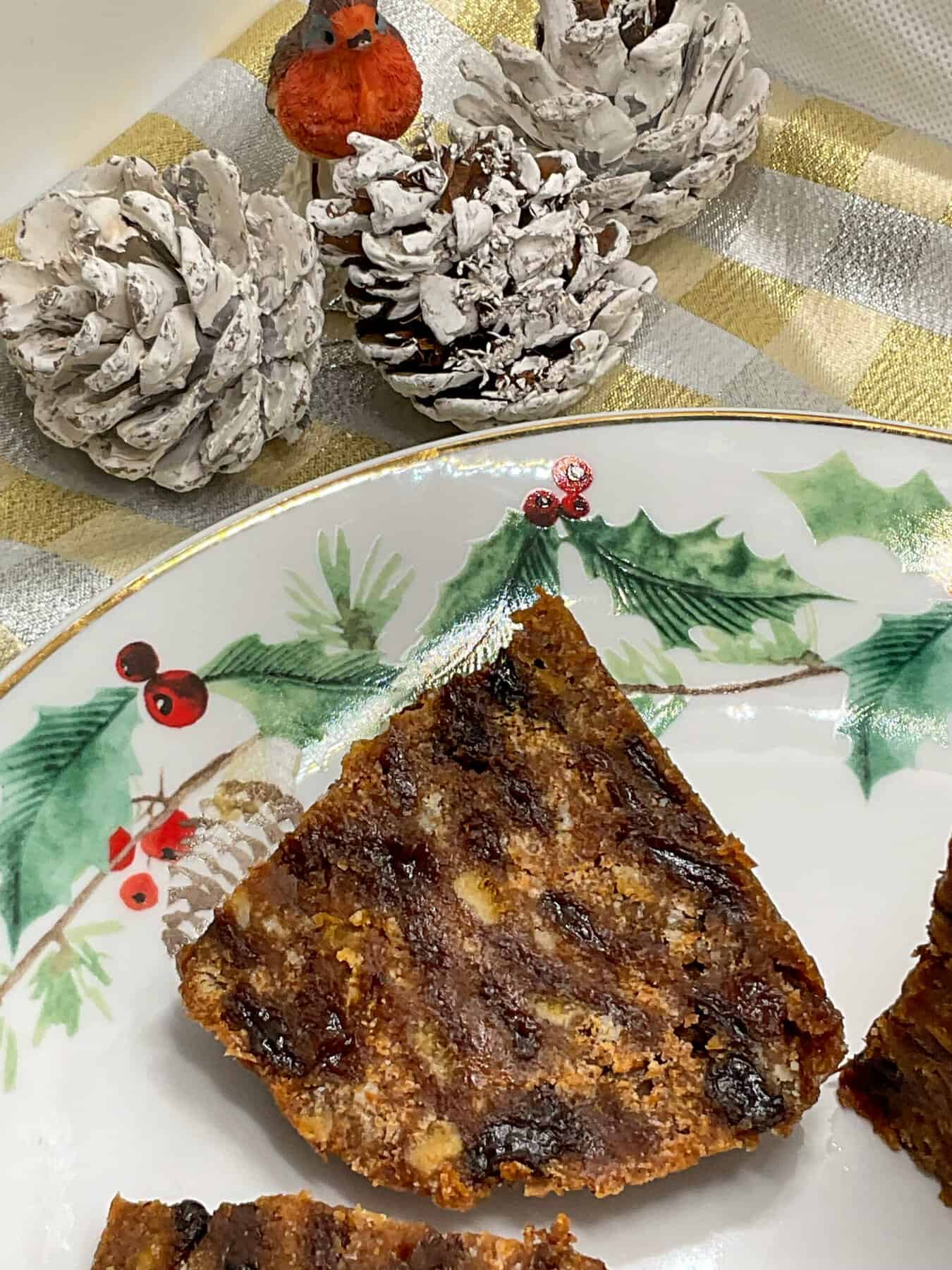 A wedge of Christmas pudding on a festive plate with holly images, frosted pine cones to side with small robin cake ornament.
