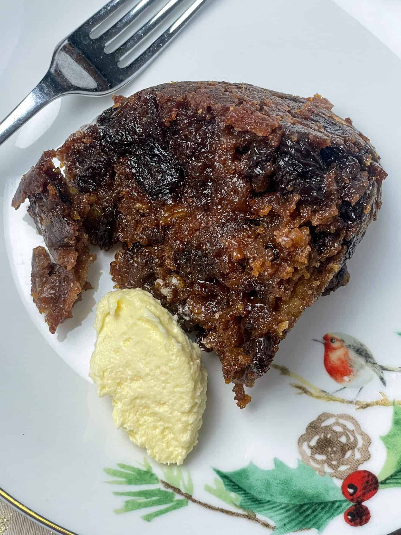 A slice of slow cooker Christmas pudding on a festive plate with a robin and tree branch image, cake fork to side of plate and a pat of brandy butter.