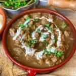 A red bowl of vegan stroganoff with pot of green veggies to side and glimpse of brown plate with brown rolls.