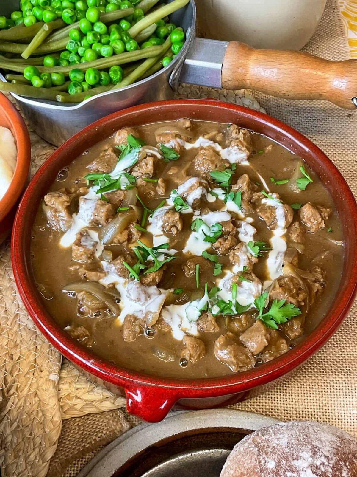 A red bowl of vegan stroganoff with pot of green veggies to side and glimpse of brown plate with brown rolls.