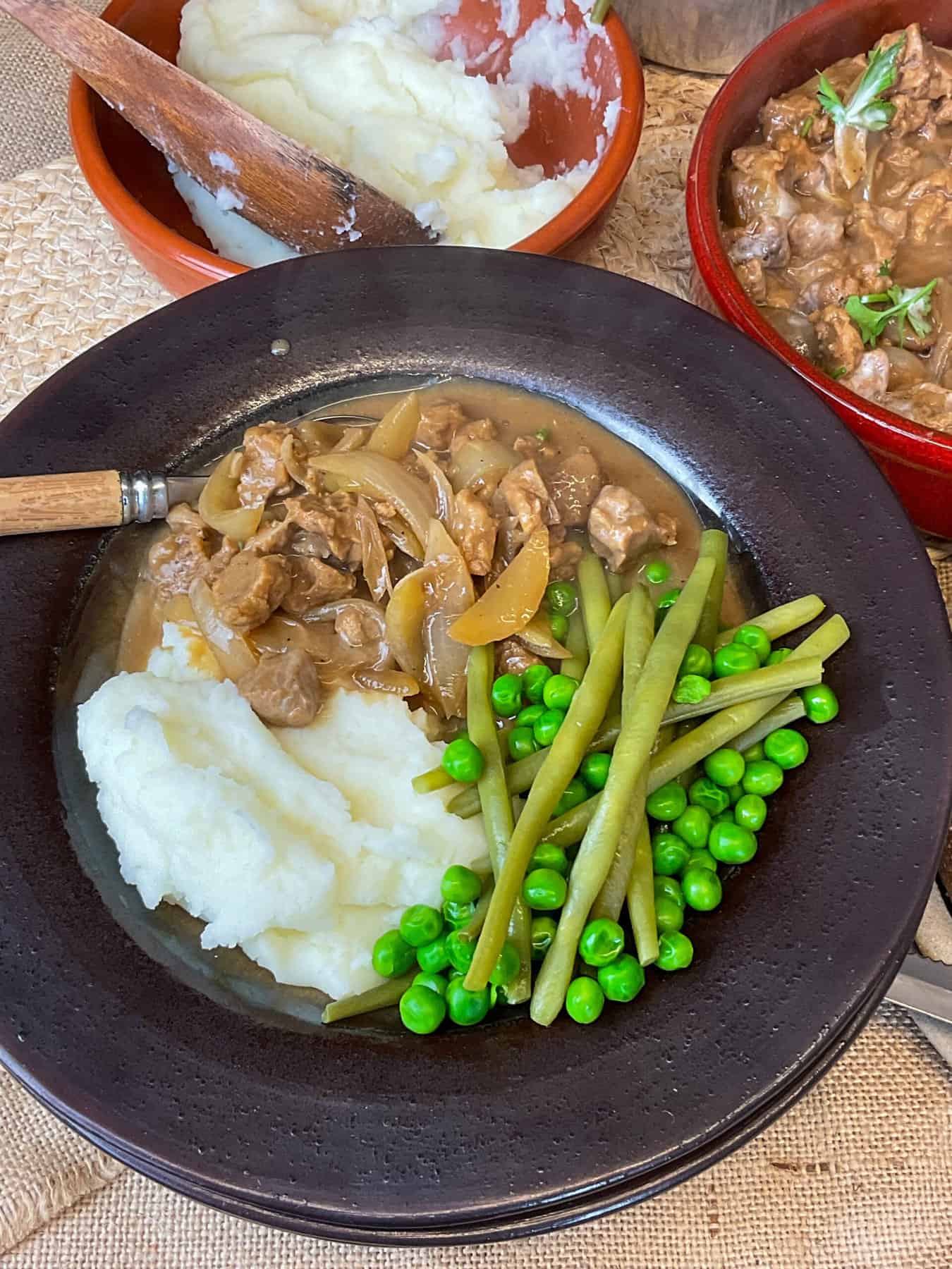 Stroganoff served in dark clay bowl with sides of mashed potato, green beans and peas, mashed potato serving bowl in background.