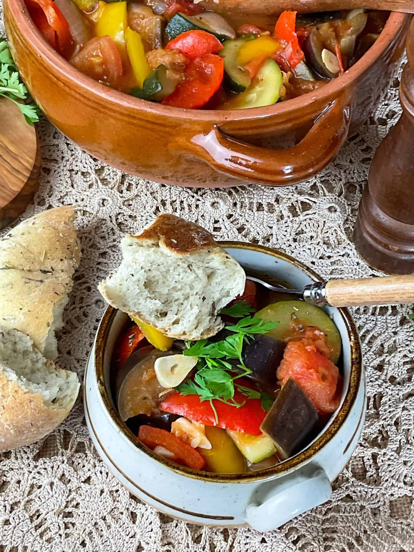 A small bowl of ratatouille with pieces of ripped bread roll at the side and a wooden handled spoon in the stew, larger serving bowl to background.