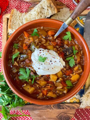 A red serving bowl filled with Hungarian pepper soup and garnished with sour cream and parsley, more parsley to side and chunks of bread to side, red table cloth background.
